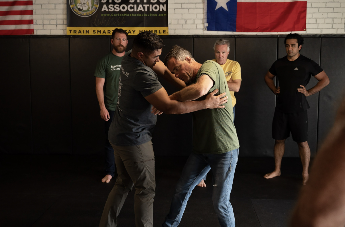 Two people in a training session, one applying a grappling hold to the other on a gym mat.